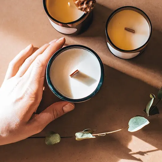 dłoń trzymająca rzemieślniczą świecę sojową oraz st&oacute;ł na kt&oacute;rym stoją dwie kolejne świece